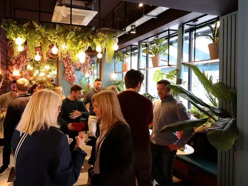 Attendees chatting beneath hanging plants and festoon lights at a Newcastle First coffee morning.